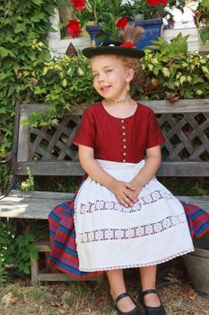 Beautiful little Bavarian girl sitting on a sunny bench in the garden in her traditional dirndl, apron and hat