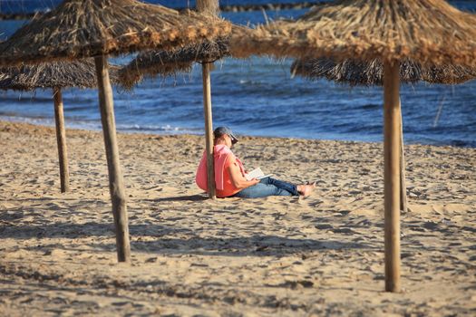Person relaxing sitting on the sand reading under a thatched beach umbrella facing the ocean