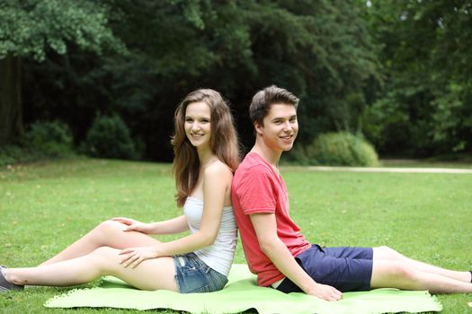 Young attractive friendly teenage couple relaxing in a lush green park sitting back to back on a rug on the grass smiling happily at the camera