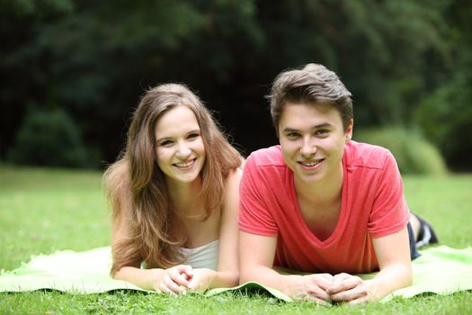 Attractive teenage boy and girl lying together on a rug on the grass facing the camera while enjoying a date in a lush green park on a summer day