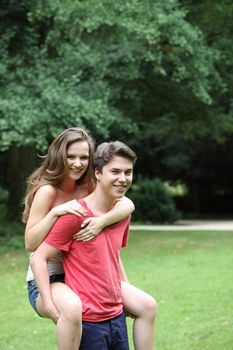 Handsome young teenage boy having fun giving his beautiful smiling girlfriend a piggyback ride in the park