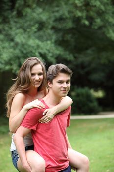 Handsome young man giving his pretty teenage girlfriend a piggyback ride outdoors in a lush green park