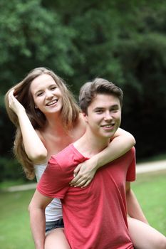 Happy laughing teenage couple having fun in the park with the boy giving the girl a piggyback ride