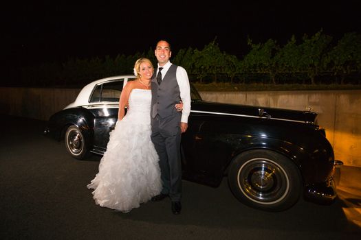 A bride and groom pose for a last photo in front of an old classic car at a vineyard at night.