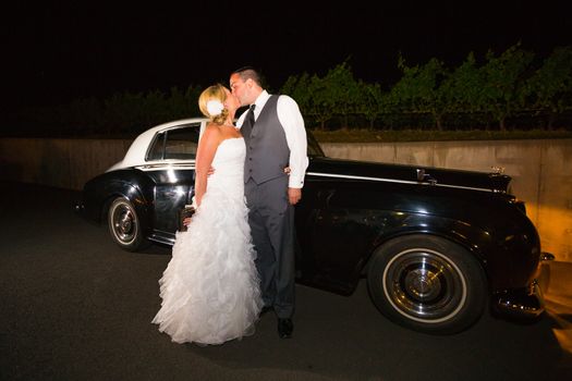 A bride and groom pose for a last photo in front of an old classic car at a vineyard at night.