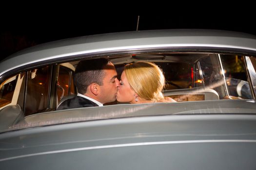 A bride and groom share a last kiss on their wedding day before leaving the reception in a classic old car at night.