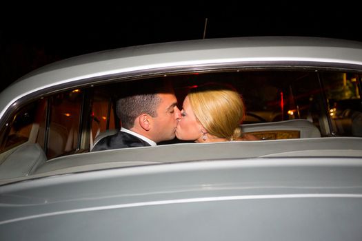 A bride and groom share a last kiss on their wedding day before leaving the reception in a classic old car at night.