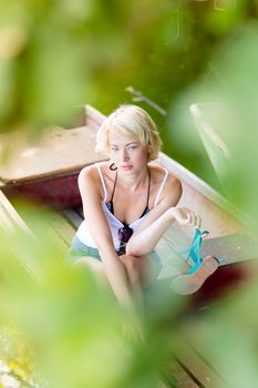 Thoughtful young blonde woman enjoying the sunny summer day on a vintage wooden boats on a lake in pure natural environment on the countryside.