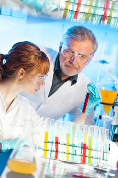 Attractive young female scientist and her senior male supervisor observing color shift of a red liquid in the glass tube in the life science research laboratory (biochemistry, genetics, forensics, microbiology..)