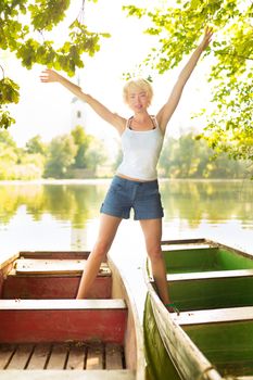 Carefree young blonde woman  arms raised and stretched enjoying the sunny summer day on a vintage wooden boats on a lake in pure natural environment on the countryside.
