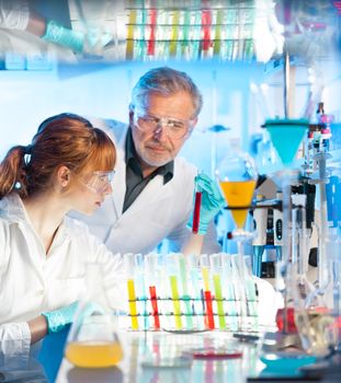 Attractive young female scientist and her senior male supervisor observing color shift of a red liquid in the glass tube in the life science research laboratory (biochemistry, genetics, forensics, microbiology..)