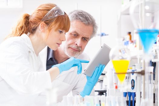 Attractive young female scientist and her senior male supervisor checking experiment protocol at tablet laptop computer in the life science research laboratory (bichemistry, genetics, forensics, microbiology..)