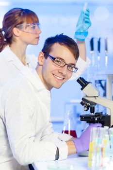 Portrait of a young male researcher microscoping in the life science (forensics, microbiology, biochemistry, genetics, oncology...)laboratory. Female asistant scientist examining blue liquid solution at the beaker in the background.
