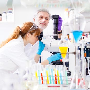 Attractive young female scientist and her senior male supervisor looking at the microscope slide in the life science research laboratory (bichemistry, genetics, forensics, microbiology..)