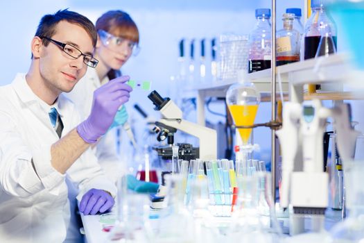 Young male researcher looking at the microscope slide in the life science (forensics, microbiology, biochemistry, genetics, oncology...)laboratory. Female asistant scientist working in the background.