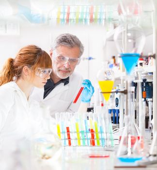 Attractive young female scientist and her senior male supervisor observing color shift of a red liquid in the glass tube in the life science research laboratory (bichemistry, genetics, forensics, microbiology..)