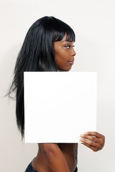 A lovely young woman holds a blank white sign while facing profile to the camera similar to a mug shot.