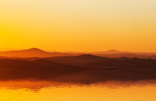 Spectacular orange sunset setting behind a low mountain range reflected in the surface of the still water in the foreground