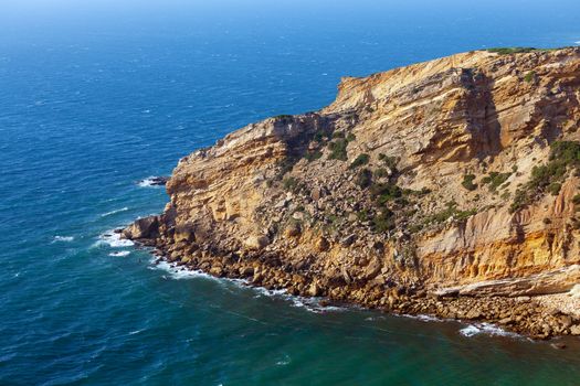 View of a rugged coastline with steep barren cliffs falling down to a rocky shoreline with waves