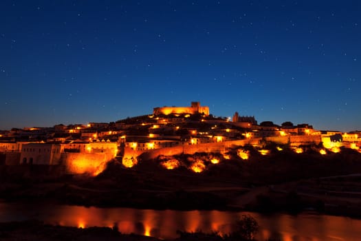 Walled town illuminated at night reflected in still water with a fortified castle on the brow of the hill overlooking the town