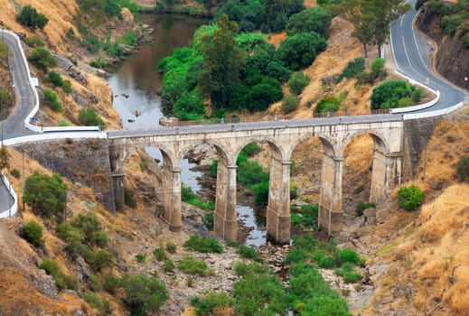 Arched road bridge with high stone arches crossing a river bed in mountainous countryside with tarred roads