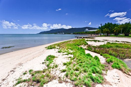 Tropical bay landscape with dune vegetation and golden sands leading to a jetty and seaside resort in the distance