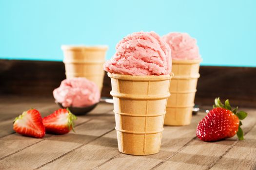 strawberry ice cream in waffle cones on wood with a scoop in background