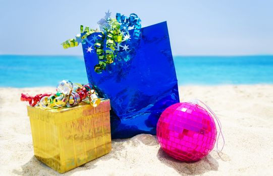 Gold gift box and blue gift bag with Christmas ball on sandy beach in sunny day- holiday concept
