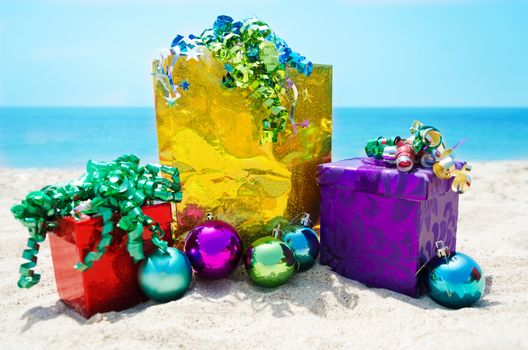 Gold gift bag and two gift boxes with Christmas balls on sandy beach in sunny day- holiday concept