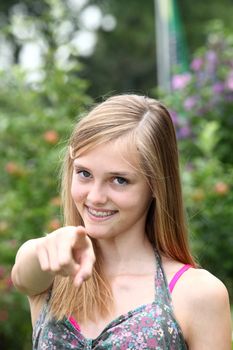 Happy teenage girl in a summer dress smiling and pointing at the camera while standing outdoors in a park or garden