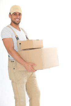 Handsome young delivery man in dungarees and a cap carrying packages and giving the camera a friendly smile isolated on white