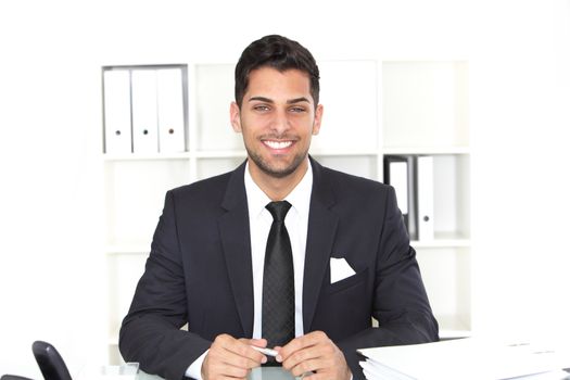 Handsome elegant smiling businessman sitting at his desk with paperwork on the desktop and binders behind him