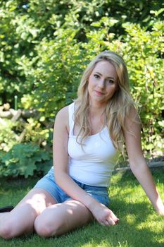 Pretty blond woman relaxing in her garden sitting on the green grass in the shade of a tree