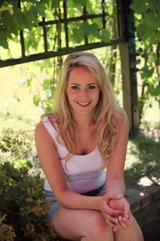 Smiling blond woman relaxing in the shade of a tree sitting on a low stone garden wall in the hot summer weather