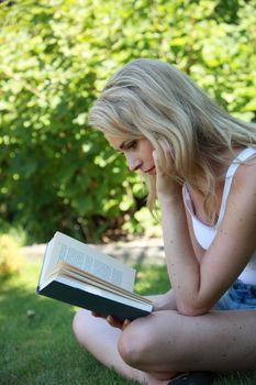 Young woman reading a book in the garden sitting sideways cross legged on the grass concentrating on the story