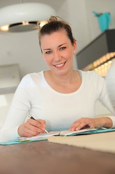 Attractive young woman working at home sitting at a table writing notes in a notebook and looking up to smile at the camera