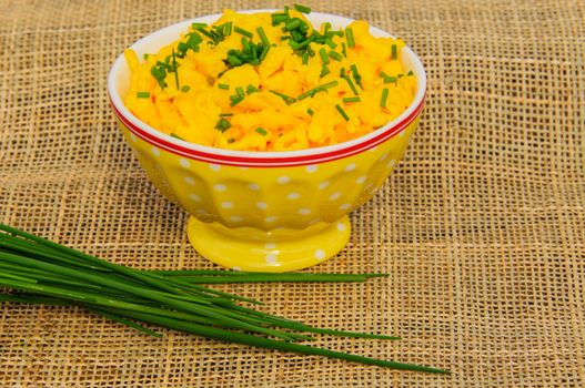 Scrambled eggs with chives in a yellow bowl on a beige napkins.  A bunch of chives lying on the napkin in front of the bowl.