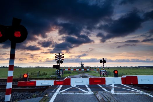 closed rail crossing with red sygnals at sunrise and windmill