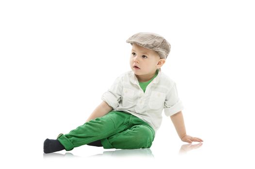Trendy little boy in a cap sitting on the studio floor looking off to the left of the frame with a solemn expression, isolated on white