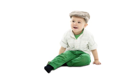 Cute dapper little boy wearing a trendy cloth hat sitting comfortably on the studio floor looking to the left of the frame with an alert amused expression, isolated on white