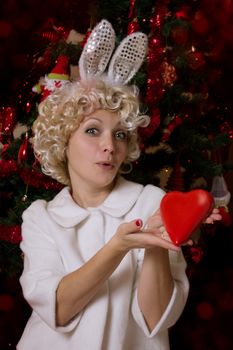 Playful woman with rabbit ears holding heart next to Christmas tree