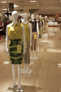 Multiple mannequins lined up in a row in a department store. The mannequins are all wearing fashionable women's clothing and are standing on a tiled floor with dramatic lighting illuminating the store. There is also a jewelry store sign in the background.