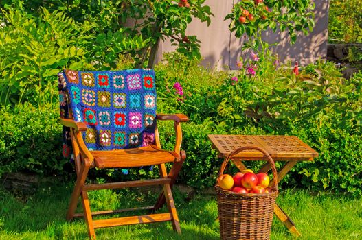 Garden furniture, patchwork carpet and a basket with apples in a garden. An apple tree in the background