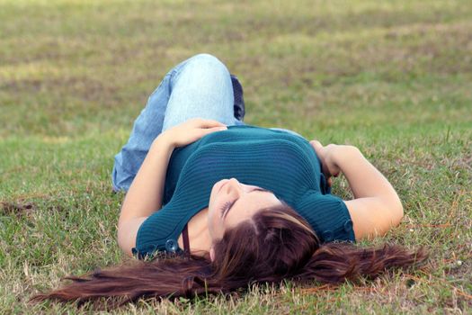 An attractive young brunette in casual wear relaxes outdoors, lying in the grass.