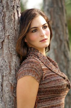 An attractive young brunette leans back against a pine tree.