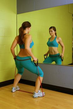 A lovely young brunette with exceptional muscle tone works out with resistance bands in front of a mirror.