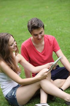 Smiling young girl and boy sitting on the green grass reading a tablet-pc as they network with their friends through social media