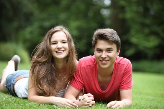 Attractive young teenage couple lying on the grass in a park holding hands and smiling at the camera