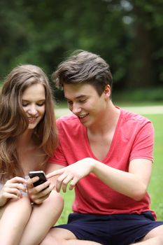 Teenage couple reading a text message on their mobile phone smiling with pleasure while sitting on grass in a park