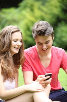 Attractive young teenage couple sitting on the lawn in a park laughing at an sms they have received on their mobile phone